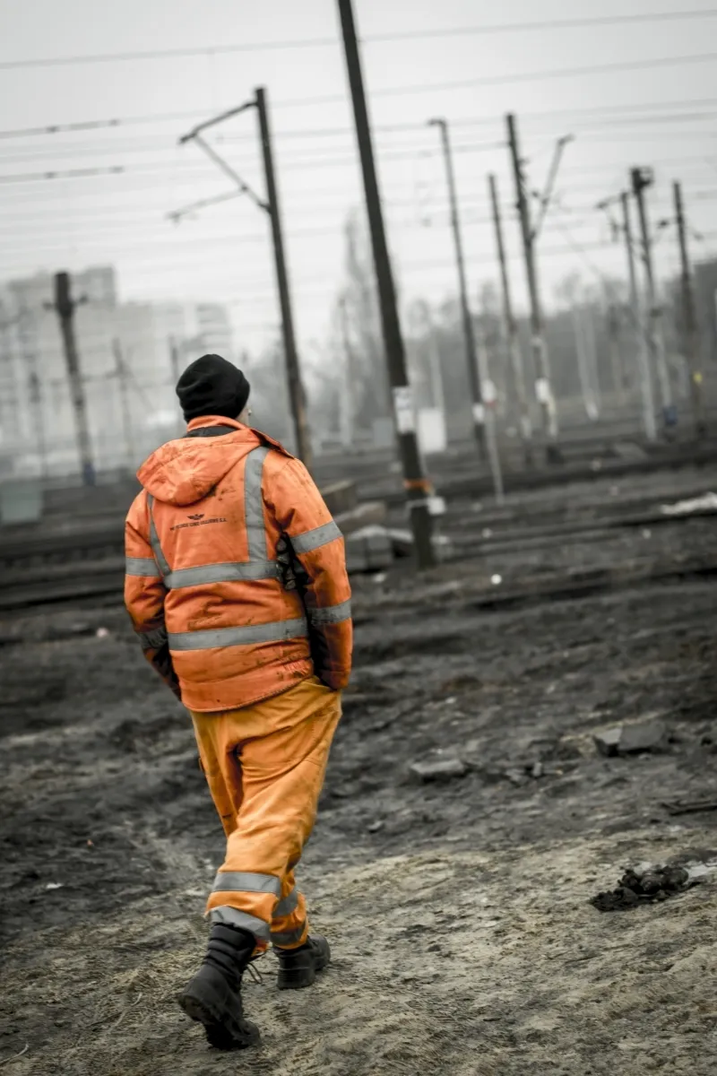 Rail worker in an orange high-visibility suit walking across muddy tracks